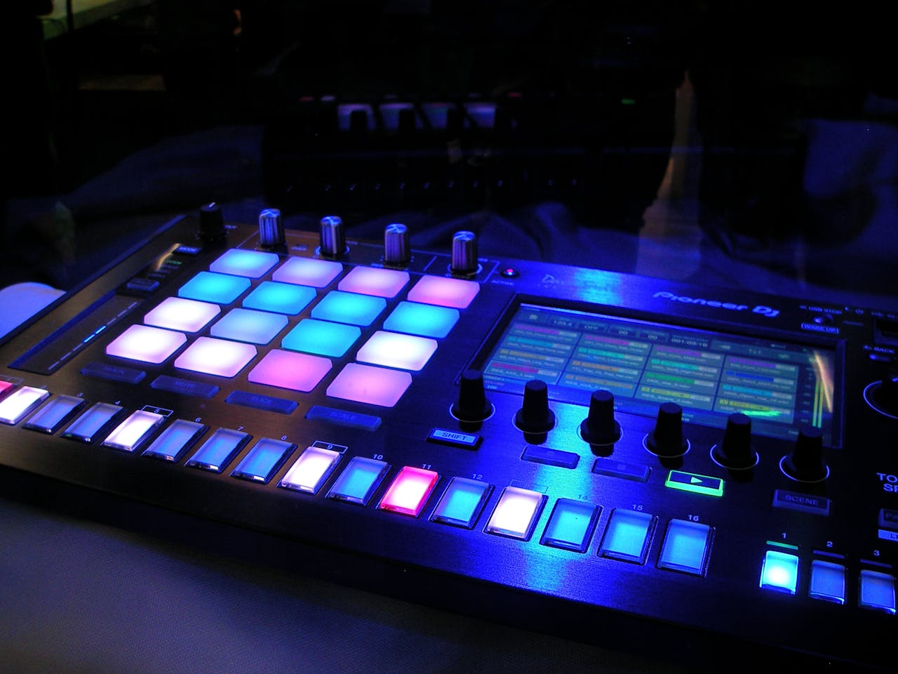services-01 Close-up of an illuminated DJ mixer with vibrant LED pads in a dark studio environment.